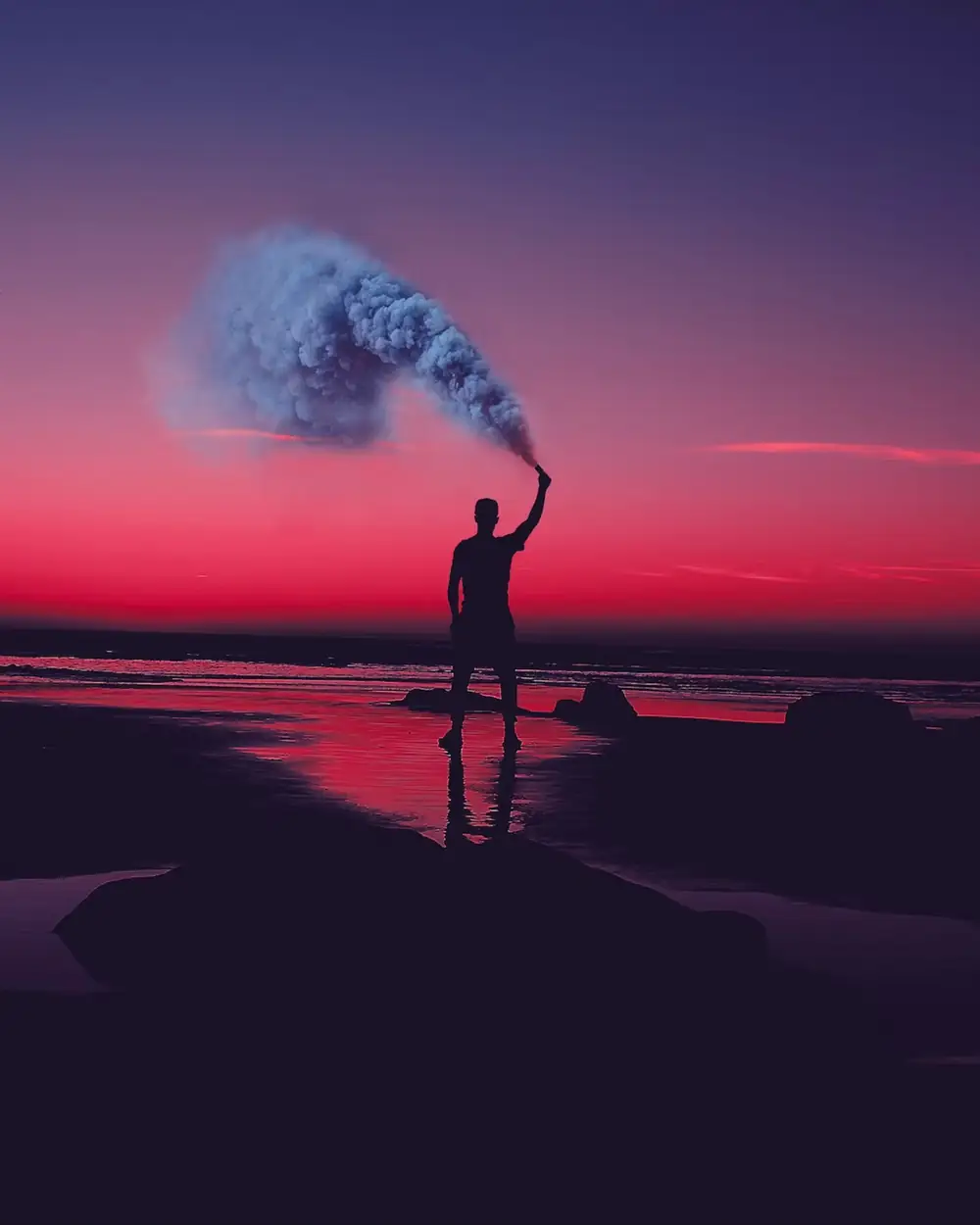 Une silhouette sur le rivage brandit un fumigène dans un ciel pourpre au coucher du soleil